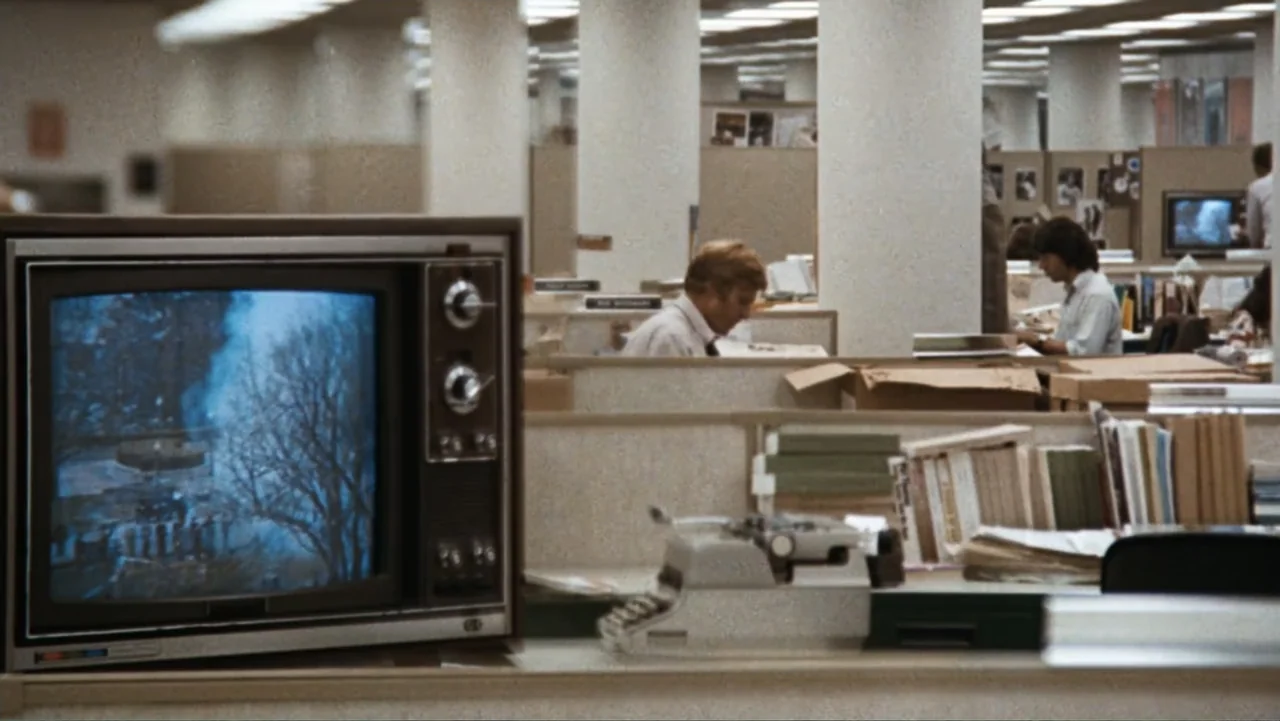 All the President's Men (1976) movie still: medium — A 1970s office with beige fabric cubicle walls and rows of structural pillars. A man sits at a…