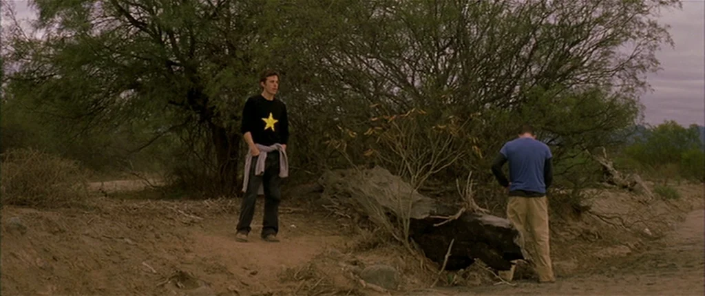 Gerry (2002) movie still: fullbody — A dry desert landscape features sparse shrubbery and a large piece of driftwood. Two young men in casual attire…