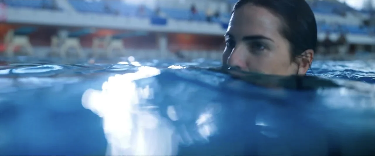 Dive (2013) movie still: closeup — A woman's face is partially submerged in the blue water of an indoor pool. The background shows blurred blue…