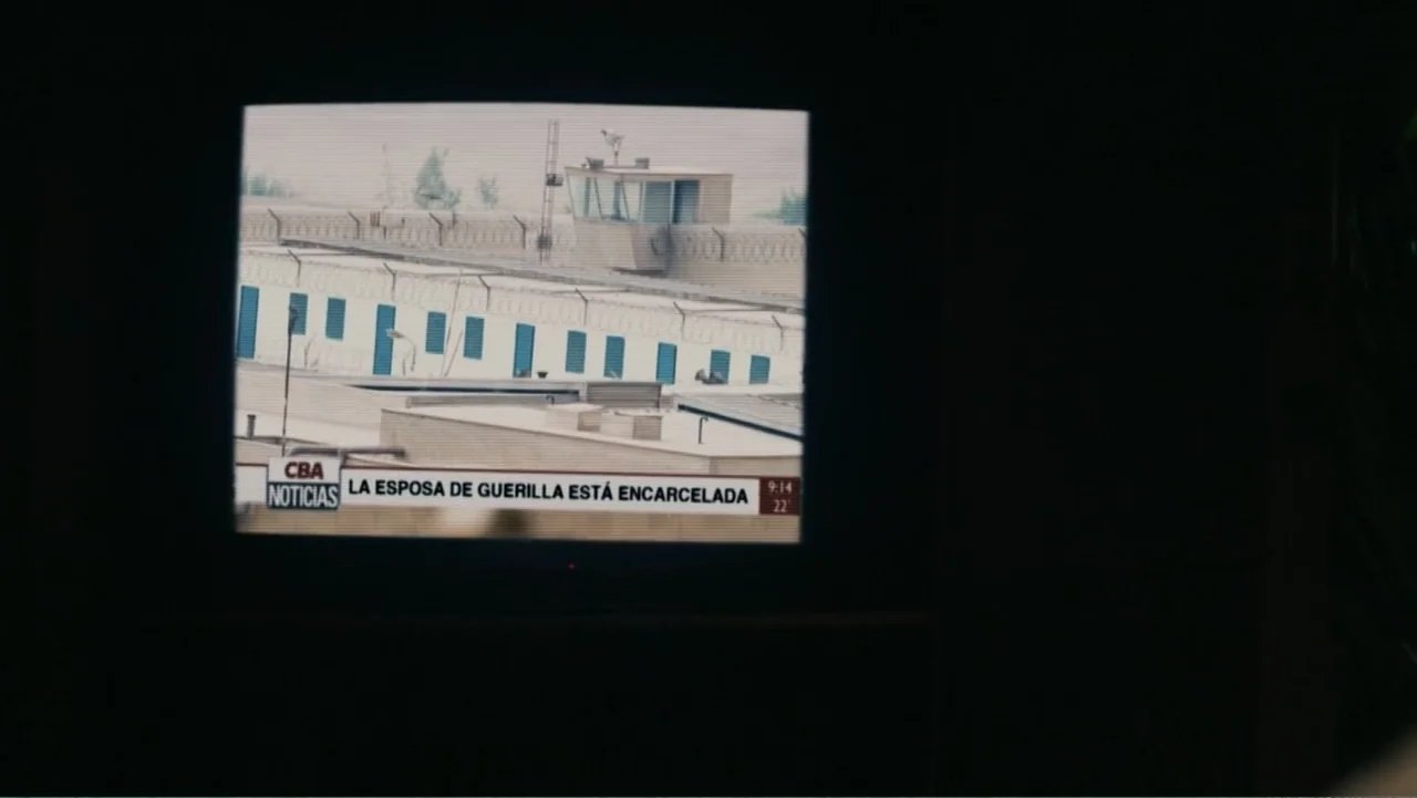 Bel Canto (2018) movie still: establishing — A television screen mounted in a dark, unlit room displays a news broadcast. The screen shows a fenced…
