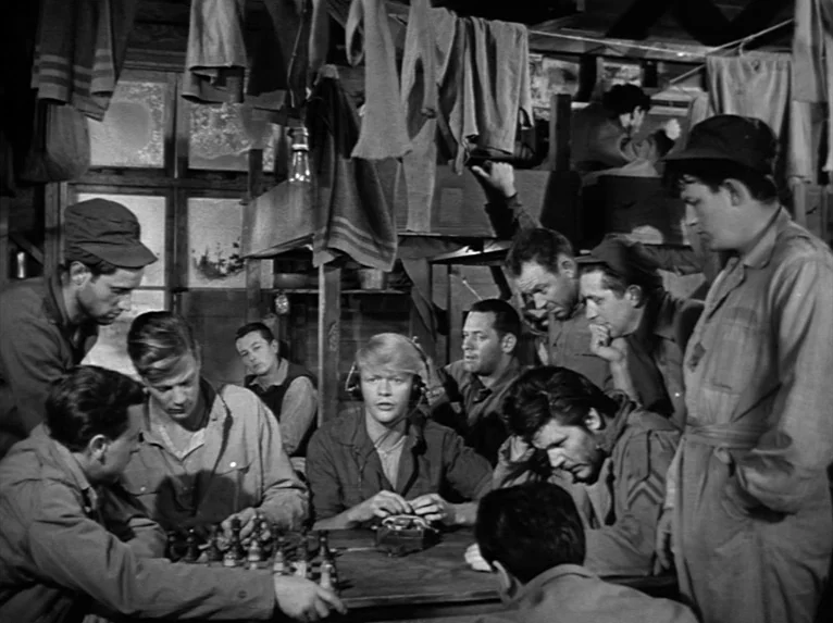 Stalag 17 (1953) movie still: medium — A crowded military bunkhouse contains men hanging laundry from low overhead rafters. A group of men plays…