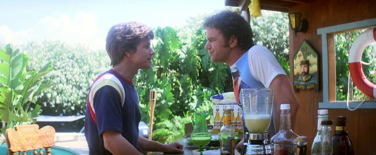 Boogie Nights movie still: medium — A sunny outdoor patio bar features lush green tropical plants in the background. Two men stand opposite each…