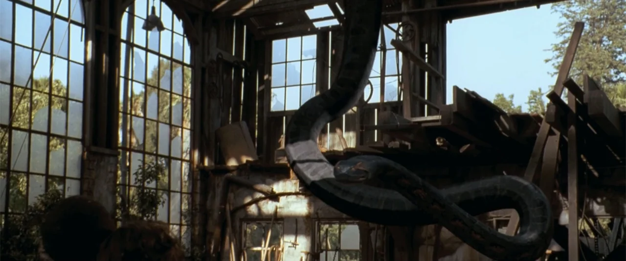 Anaconda (2025) movie still: wide — A dilapidated greenhouse features cracked glass panels and wooden structural remains. A giant snake coils…