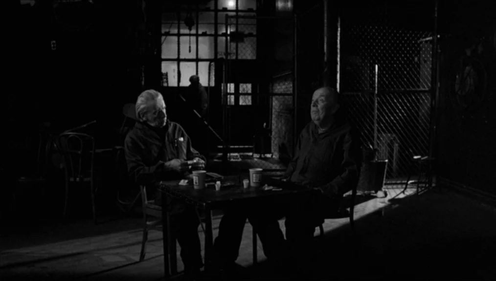 Coffee and Cigarettes (2003) movie still: medium — A dimly lit room with chain-link fencing and dark industrial walls in the background. Two older…