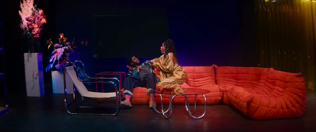 Rye Lane (2023) movie still: wide — A spacious room features a bright orange modular sofa set against a dark blue wall. A woman kneels on the sofa…