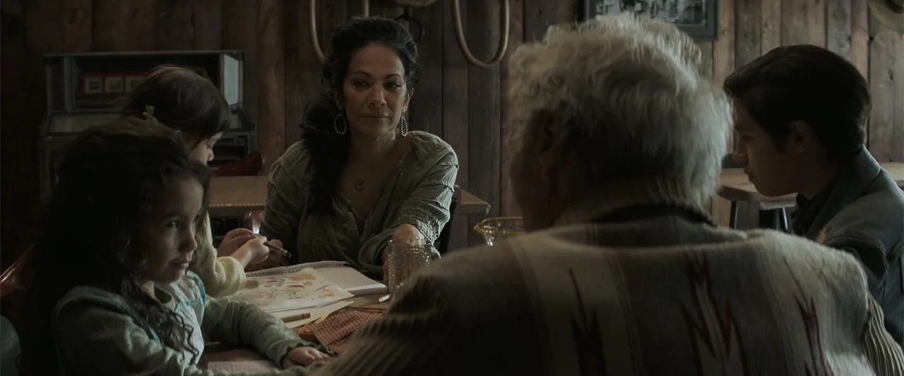 Cry Macho (2021) movie still: medium — A rustic wooden room features vertical plank walls and a table where people gather. A woman sits with two…