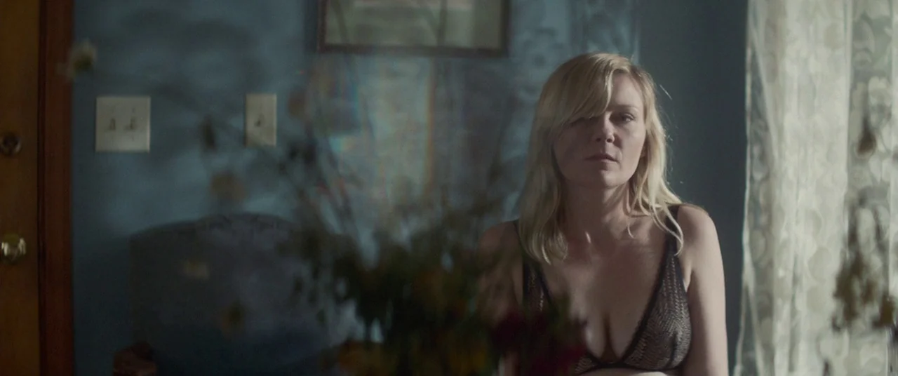 Woodshock (2017) movie still: medium — A dimly lit room with dark blue walls features a window covered by lace curtains. A blonde woman sits in the…