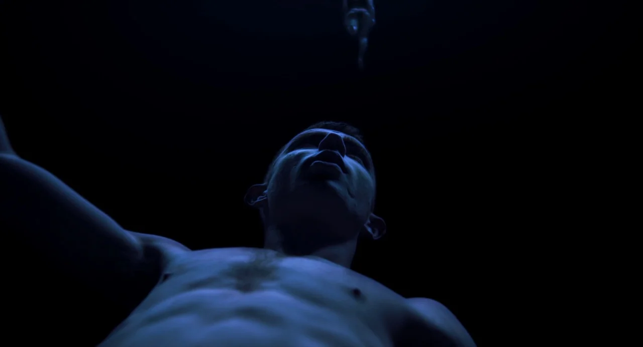Under The Skin (2013) movie still: closeup — A dark, empty space surrounds a shirtless man shown from a low angle. He looks upward with his mouth…