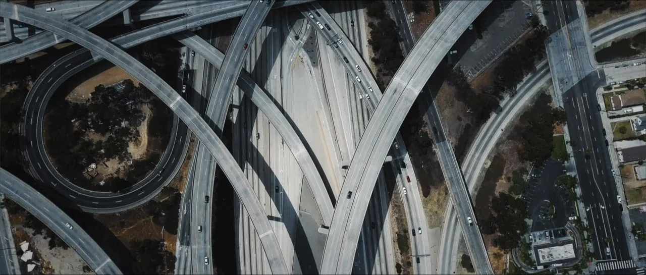 Square One (2019) movie still: establishing — An aerial view of a multi-level highway interchange with complex intersecting concrete bridges.…