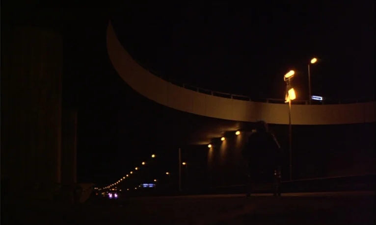 Spetters (1980) movie still: wide — A dark, curved highway overpass stretches into the distance under artificial amber street lighting. A solitary…