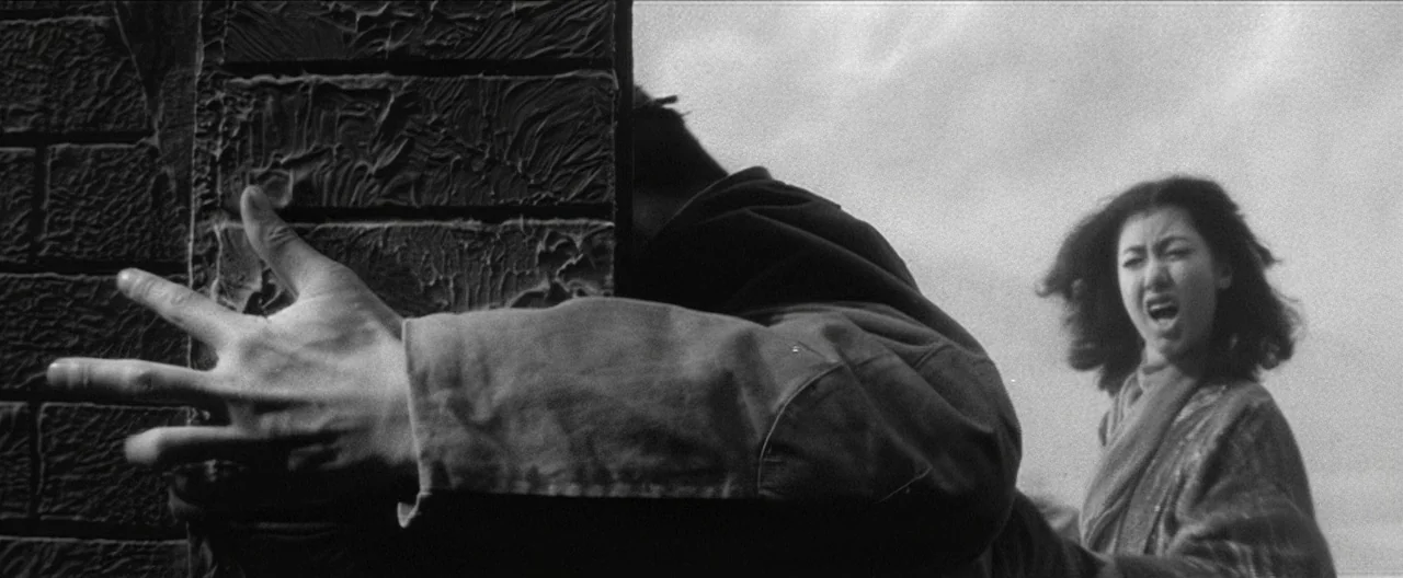Story of a Prostitute (1965) movie still: medium — A rooftop setting features a textured brick column and overcast sky. A woman yells toward a man…