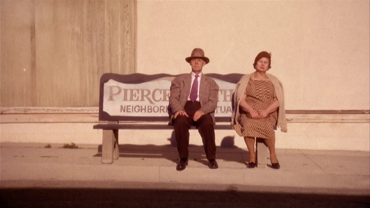 The Day of the Locust (1975) movie still: fullbody — A man and a woman sit together on a wooden bench against a wall. The bench displays the words…