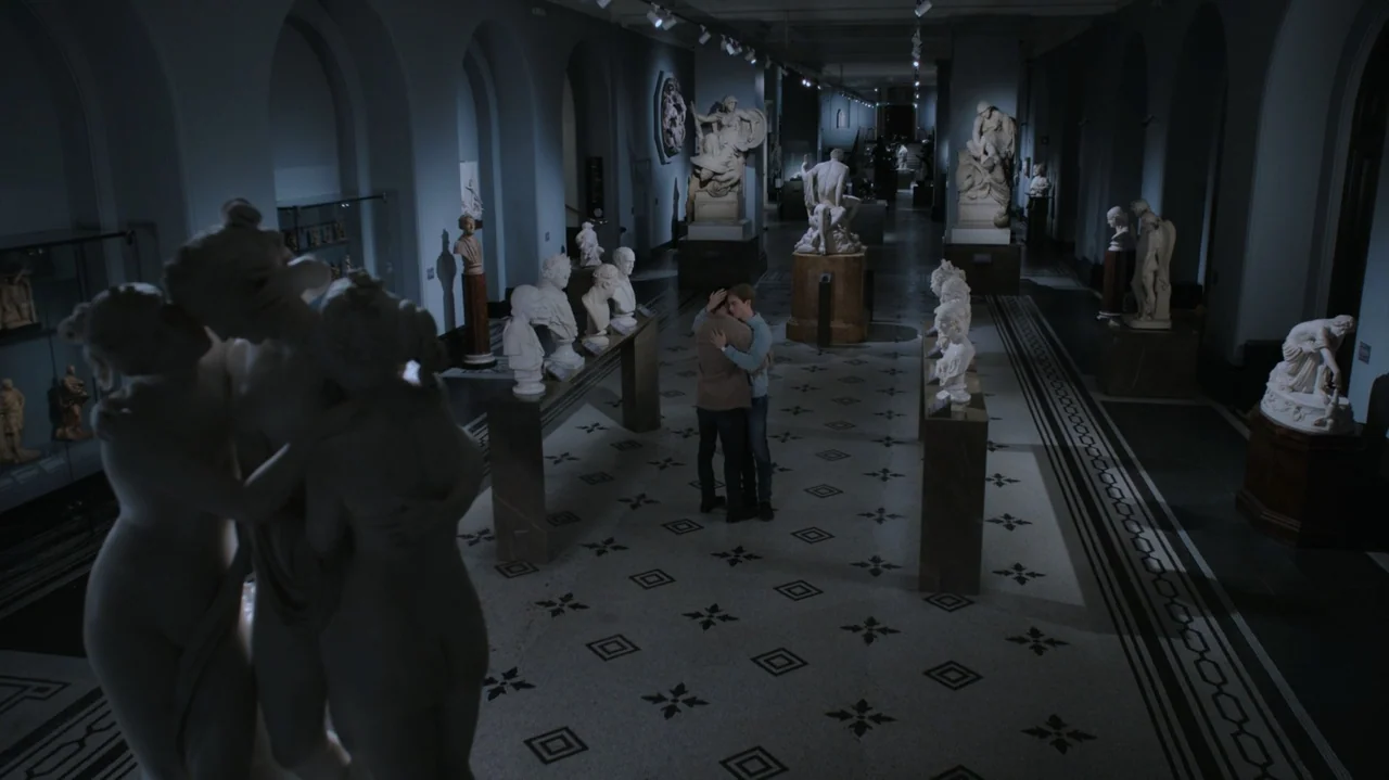 Red, White & Royal Blue (2023) movie still: wide — A spacious museum hall features rows of white marble busts and sculptures on pedestals. Two men…