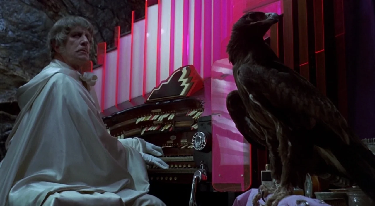 Dr. Phibes Rises Again (1972) movie still: medium — A dark rocky cave contains a large pipe organ with backlit pink glass panels. A man in a white…