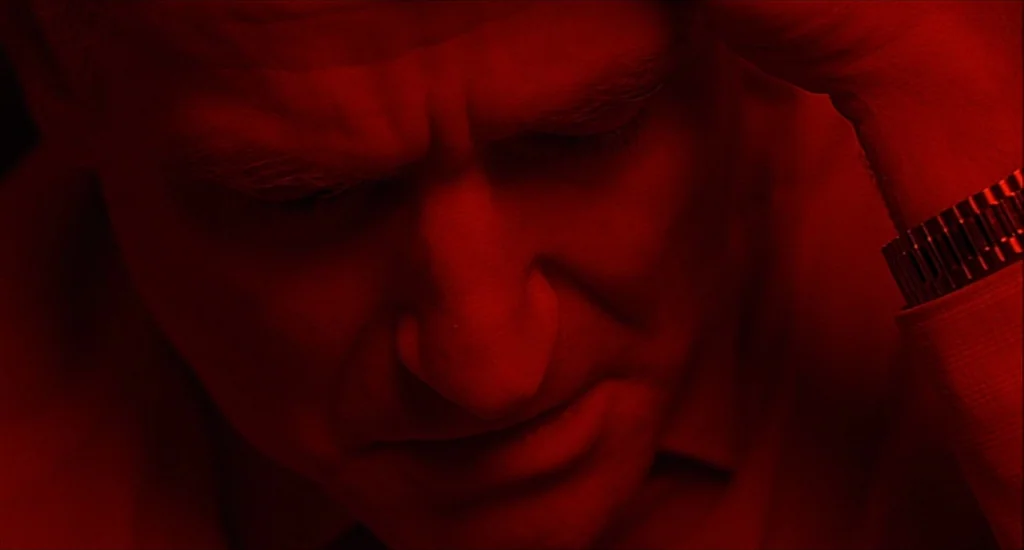 One Hour Photo (2002) movie still: closeup — The man sits in a dark space completely bathed in monochromatic red light. His head is bowed with his…