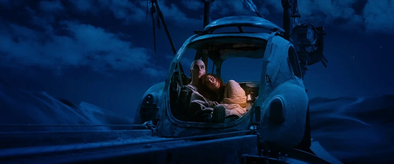 Mad Max: Fury Road (2015) movie still: wide — A vast, dark blue desert landscape stretches under a starry night sky. Two individuals huddle together…