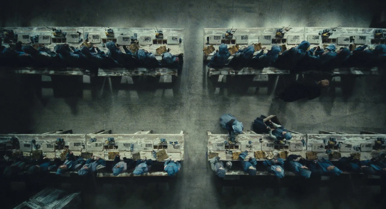 Les Misérables (2012) movie still: establishing — A top-down view shows rows of long wooden tables in an industrial factory setting. Numerous…