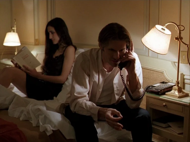 For Ever Mozart (1996) movie still: medium — A dimly lit hotel room features a double bed with two occupants in the evening. The man speaks on a…