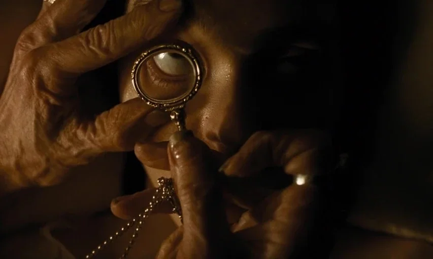 Nosferatu (2024) movie still: closeup — A dark indoor setting features a person holding a magnifying glass against their face. The lens enlarges one…