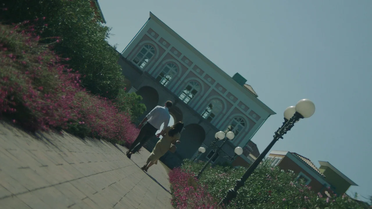 Infinity Pool (2023) movie still: fullbody — A paved path lined with pink flowers leads toward a multi-story building facade. A young man and woman…