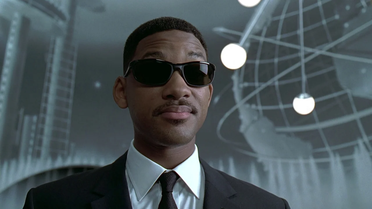 Men In Black (1997) movie still: closeup — A man stands before a large metal sculpture of a globe at night. The man in a black suit looks directly…