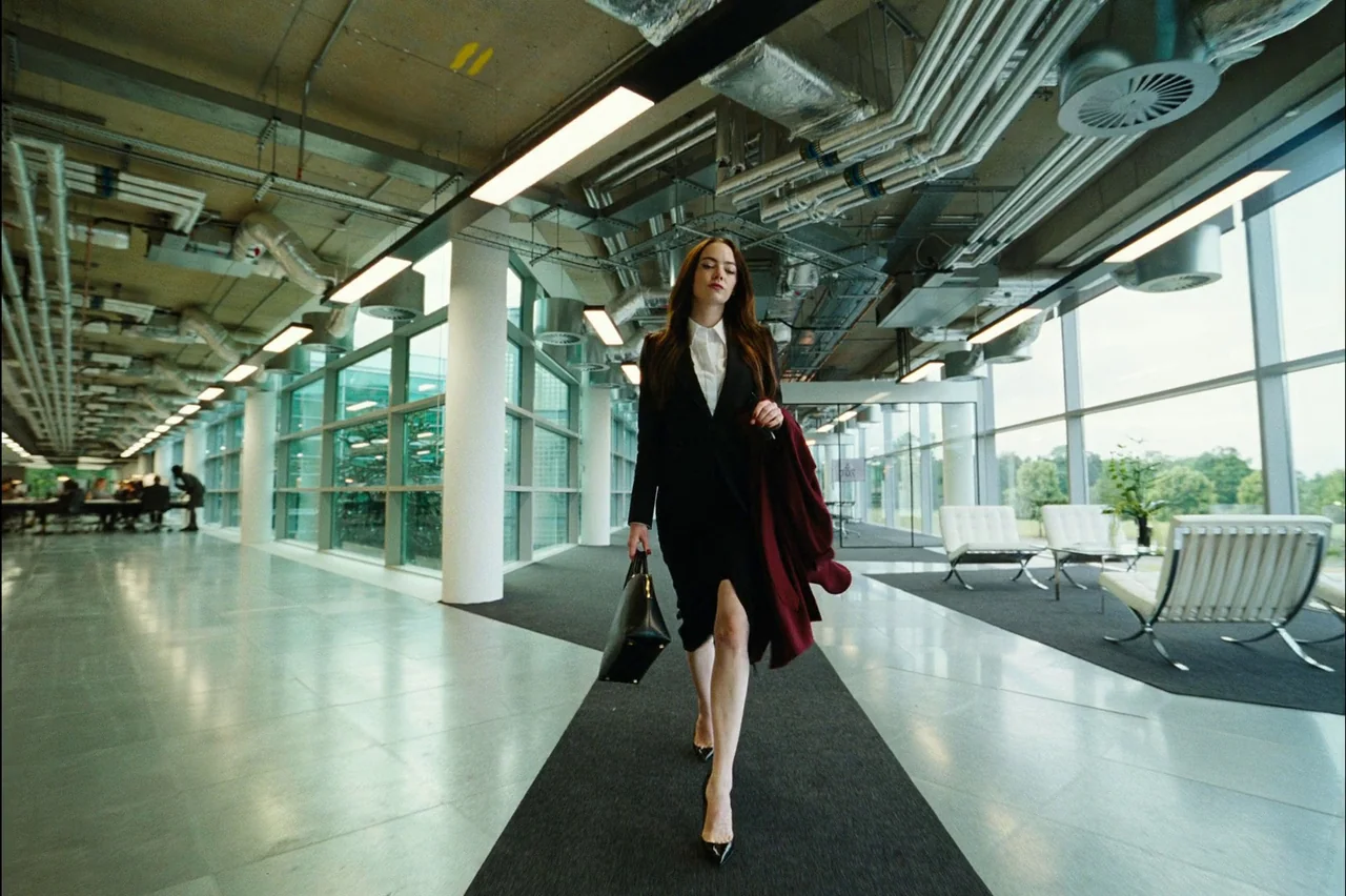 Bugonia (2025) movie still: fullbody — A spacious office interior features exposed industrial pipes overhead and long glass-walled rooms. A woman in…