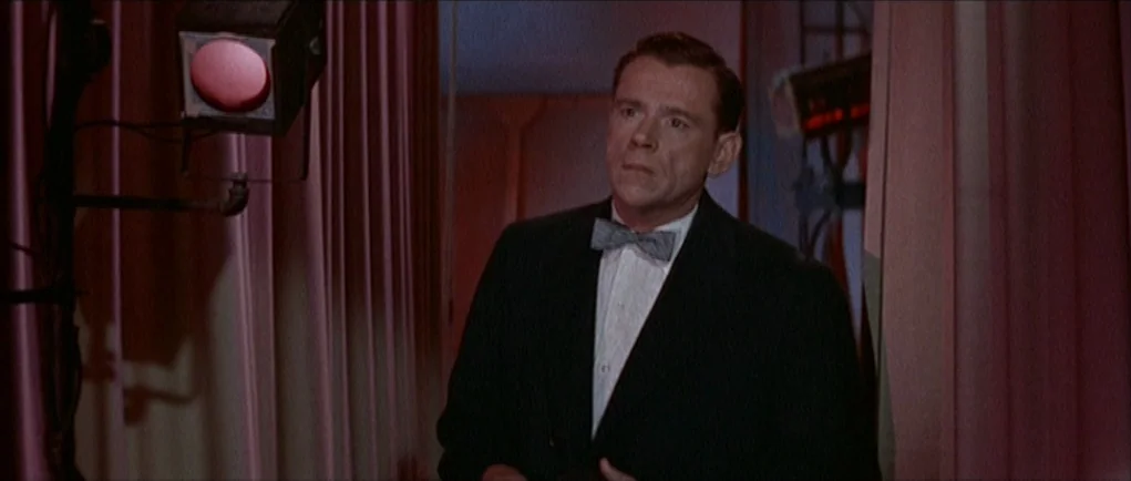 The Girl Can’t Help It (1956) movie still: medium — A man in formal wear stands near a studio light on a theatrical set. Heavy pink curtains form…