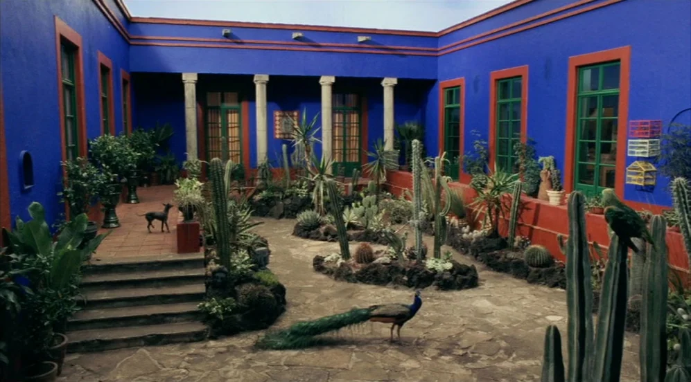 Frida (2002) movie still: establishing — A spacious courtyard features bright blue walls with terracotta trim and stone floors. Cacti and potted…