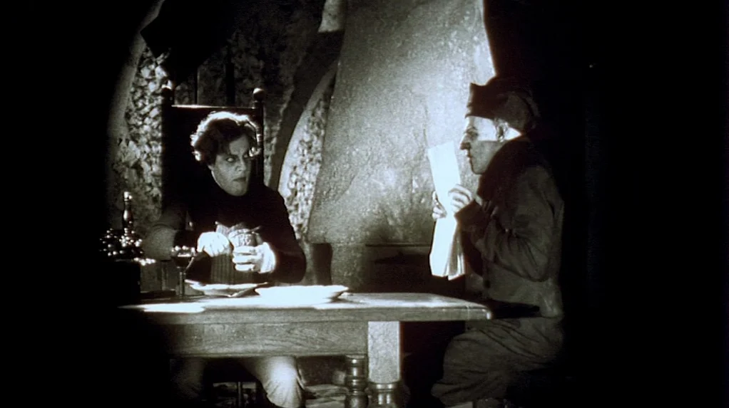 Shadow of the Vampire (2000) movie still: medium — A stone-walled interior with a heavy fireplace behind a wooden table. Two men sit opposite each…