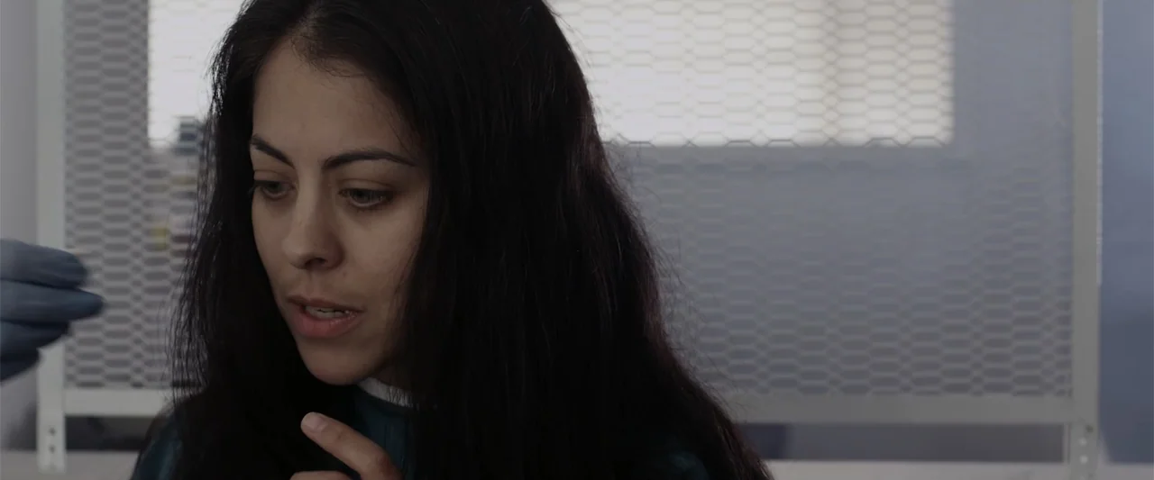 Mermaid Down (2019) movie still: closeup — A clinical space with a white metal mesh partition visible in the blurred background. A young woman in…