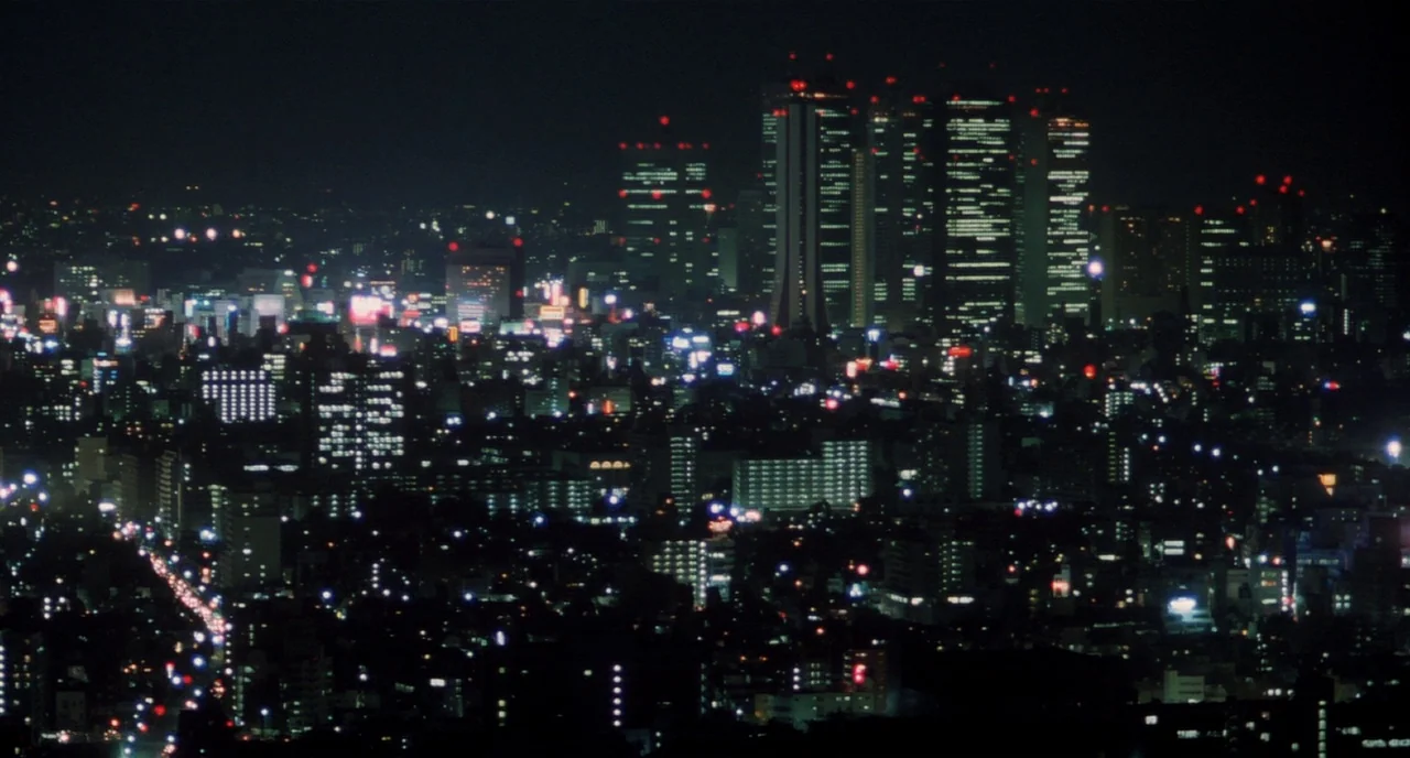 Tampopo (1985) movie still: establishing — A dense urban sprawl stretches across the horizon at night under a dark sky. Tall skyscrapers with grid…