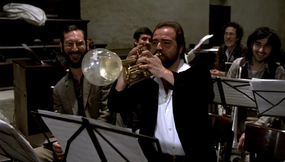Orchestra Rehearsal (1978) movie still: medium — A dimly lit rehearsal space features musicians seated with music stands in a cluster. A bearded man…