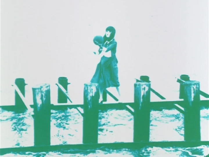 Labyrinth Tale (1975) movie still: fullbody — A woman stands atop a wooden pier pilings structure overlooking the water. She holds a round ball-like…