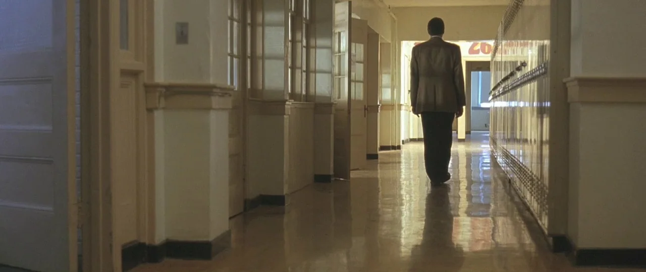 In the Bedroom (2001) movie still: fullbody — A long school corridor with lockers on one side and wooden classroom doors. A man walks away from the…