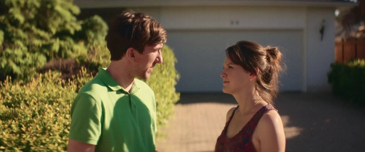 Room For Rent (2017) movie still: medium — A suburban residential driveway with a house and garage in the background. A man and a woman stand close…
