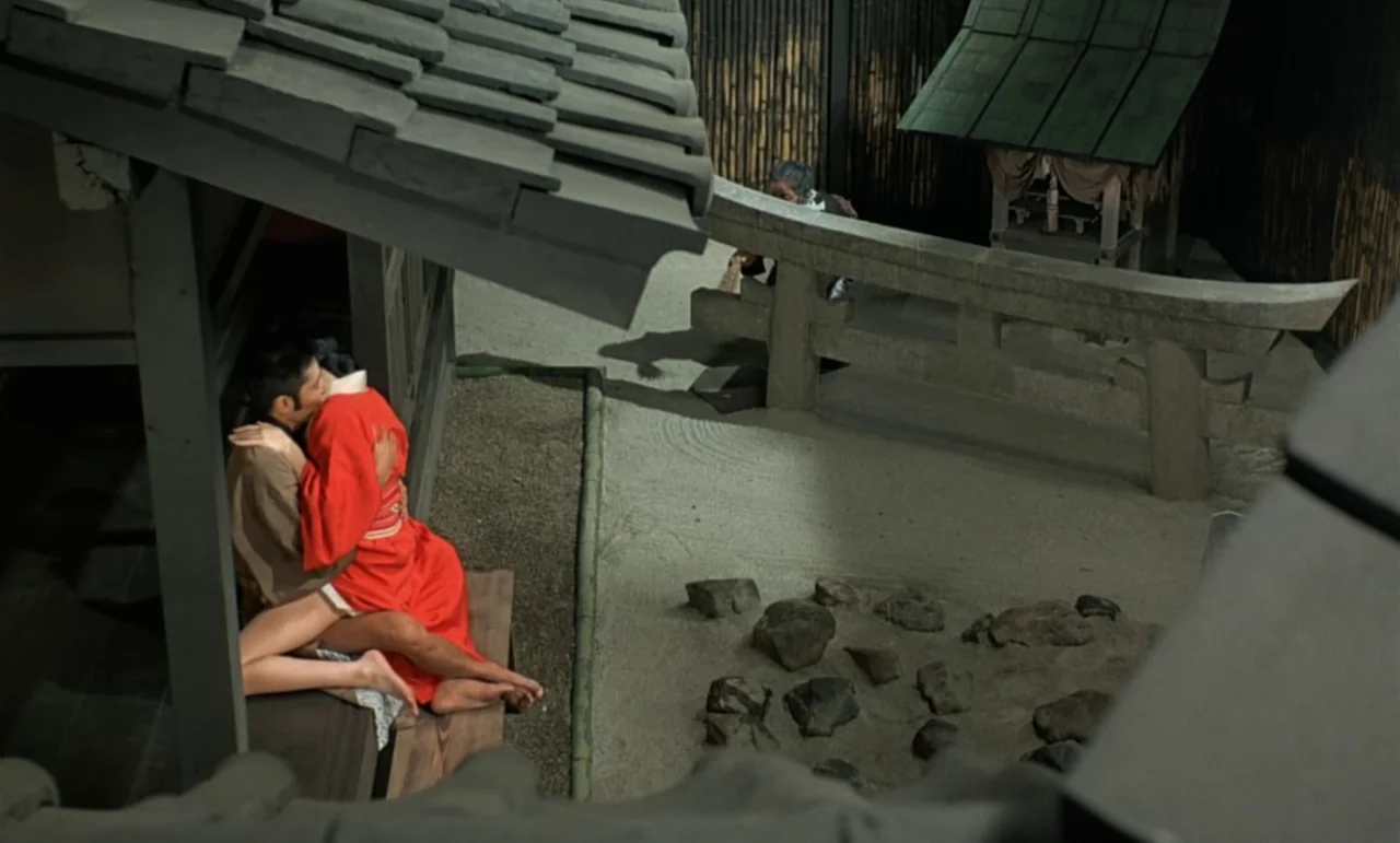 In The Realm of the Senses (1976) movie still: wide — A traditional Zen stone garden with a gray tiled roof covering a wooden porch. A couple sits…