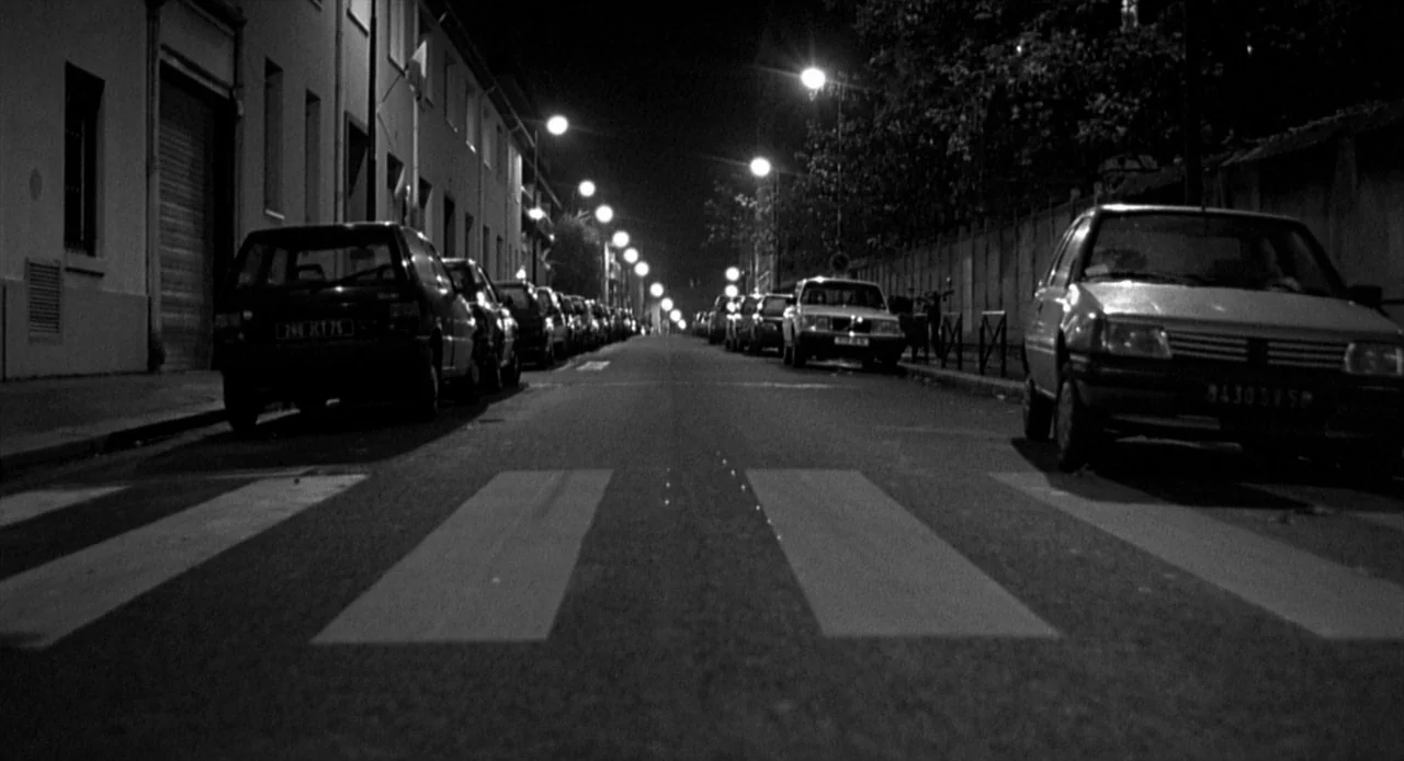 La Haine (1995) movie still: establishing — A narrow city street at night is flanked by residential buildings and parked vehicles. The perspective…