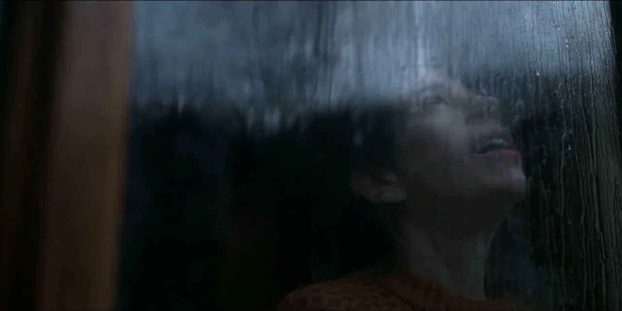 Bring Her Back (2025) movie still: closeup — A close-up view through a rain-covered window pane into a dark interior space. A person stands near the…