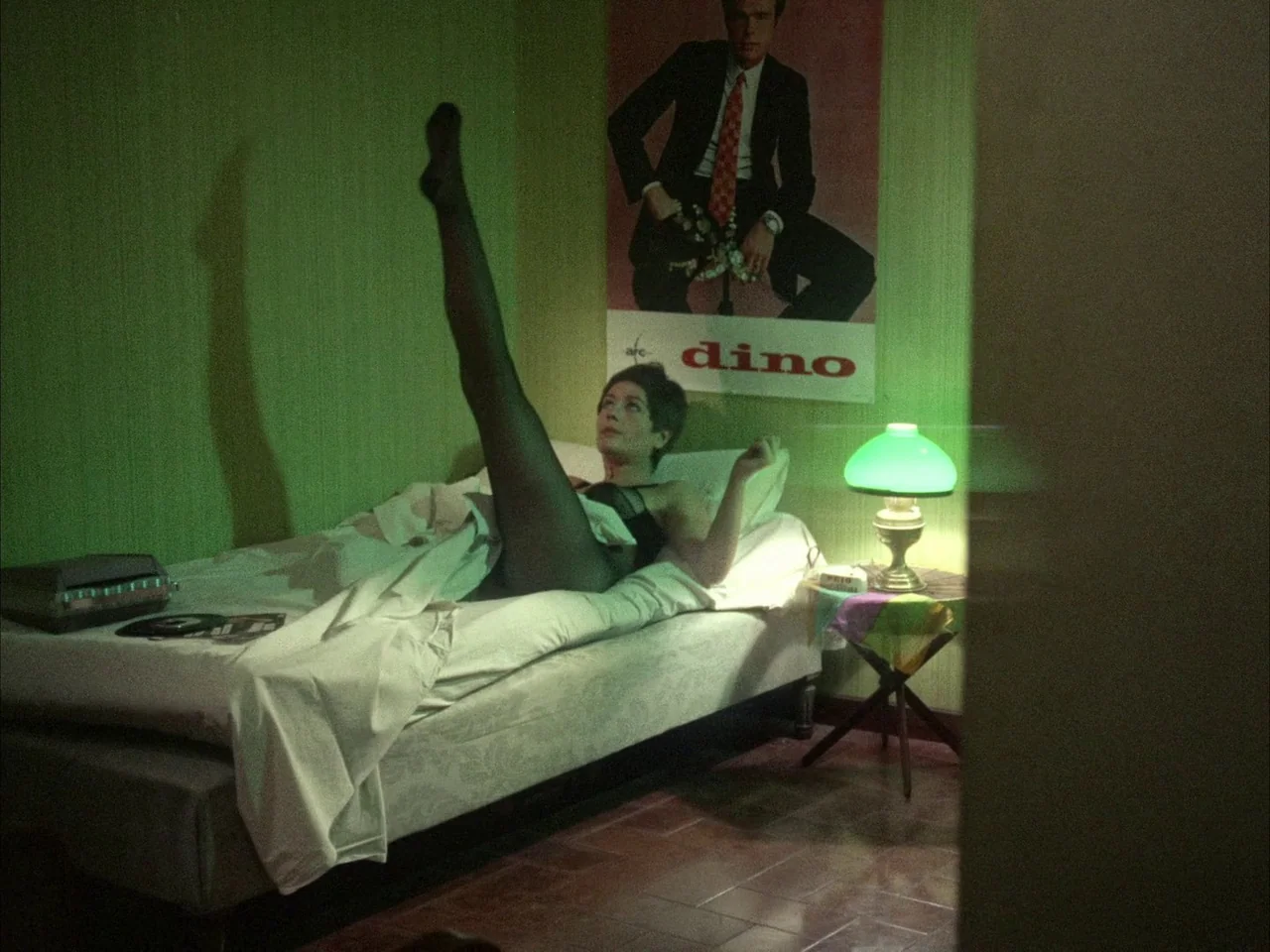 Dillinger is Dead (1969) movie still: medium — A green-walled hotel room contains a bed and a vintage bedside table lamp. A woman wearing a leotard…