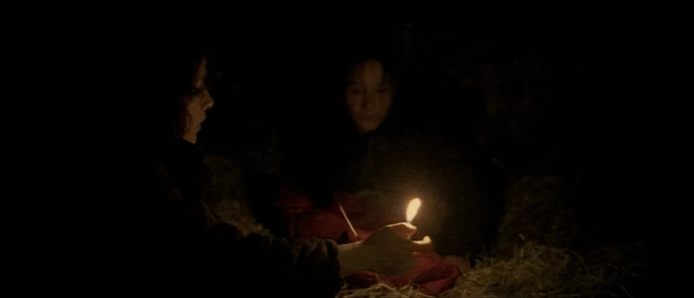 Time Of The Wolf (2003) movie still: medium — Two figures are situated inside a dark hayloft surrounded by dry straw. One figure holds a small,…