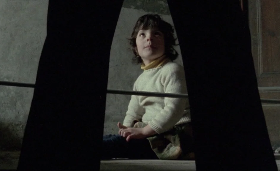 The Kingdom of Naples (1978) movie still: medium — A child sits on a wooden floor in a dimly lit room. The child looks upward toward a person…