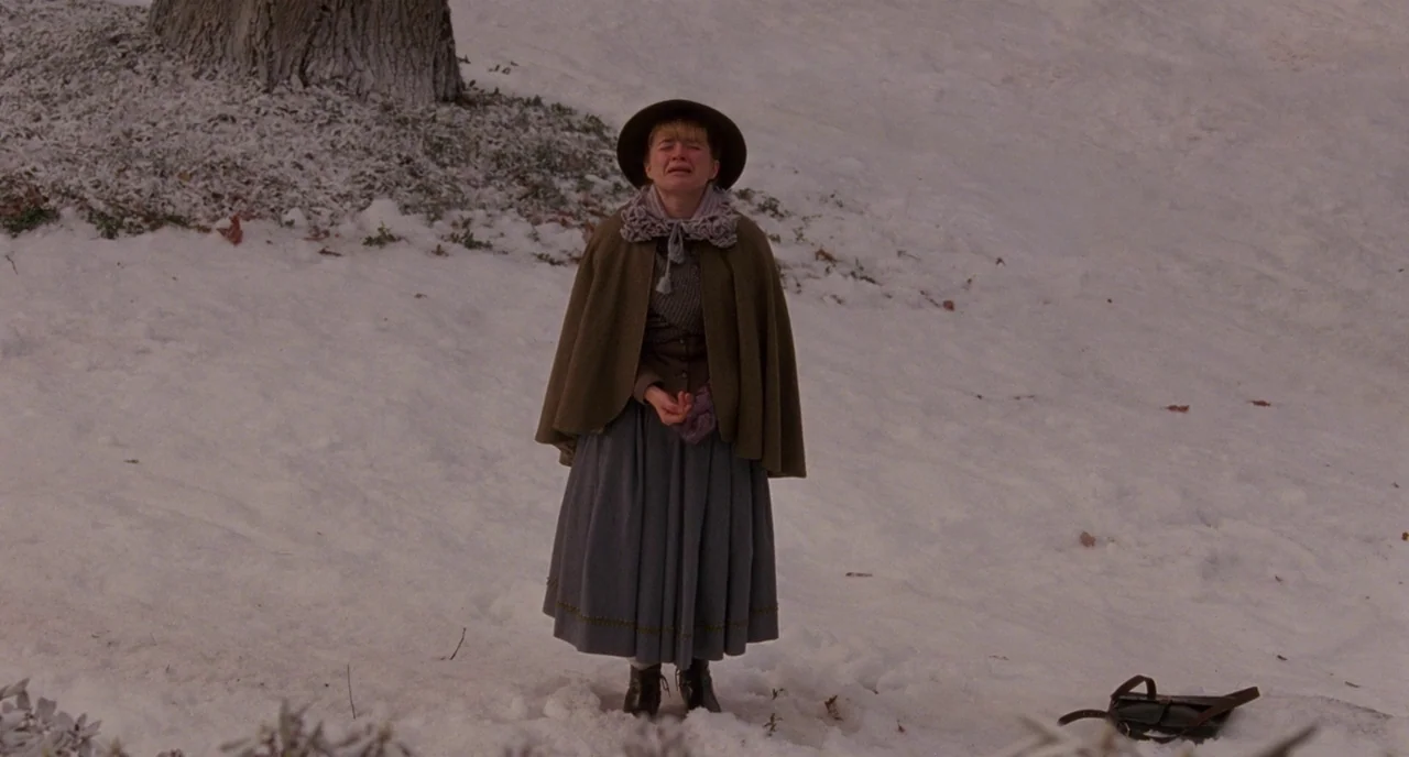 Little Women (2019) movie still: fullbody — A snowy hillside with a single large tree trunk on the left side. A woman stands in the snow with her…