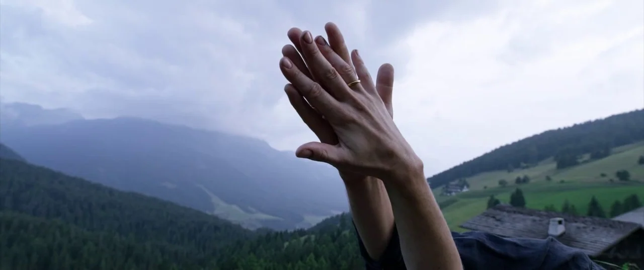 A Hidden Life (2019) movie still: closeup — A mountain landscape under a cloudy, overcast sky stretches into the distance. Two human hands, one with…