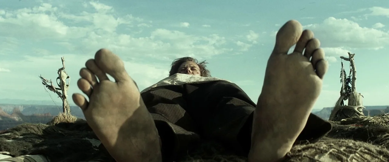 The Lone Ranger (2013) movie still: closeup — A man lies flat on a dusty, rocky desert plateau under a bright sky. Two small wooden totems with…