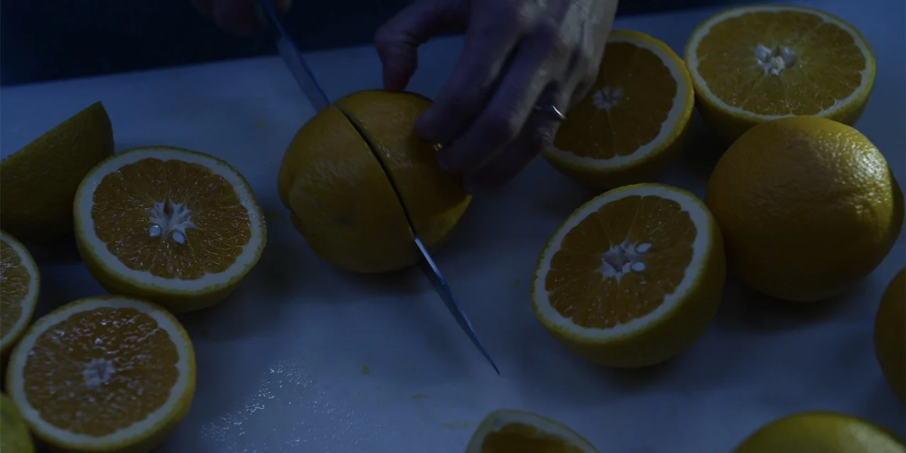About Fate (2022) movie still: closeup — A white cutting board is covered in halved oranges under dim light. A silver knife divides an orange half…