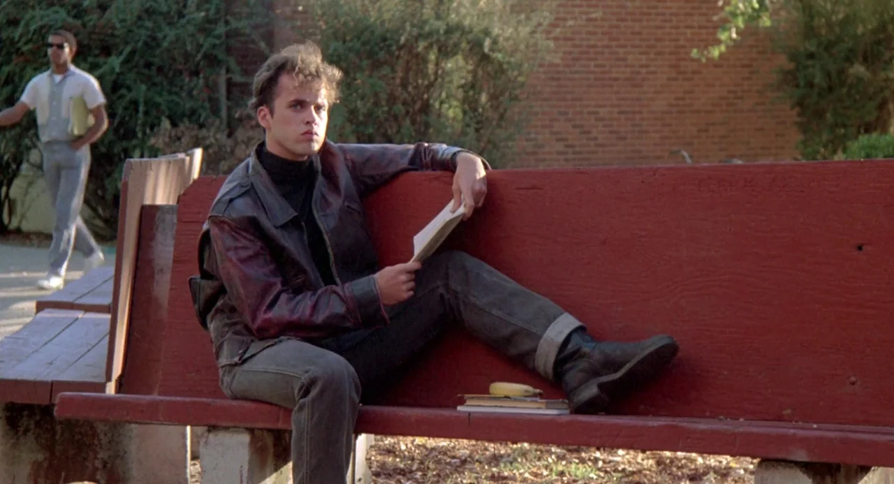 Peggy Sue Got Married (1986) movie still: medium — A person sits on a painted red bench on an outdoor campus grounds. A book rests on his lap while…