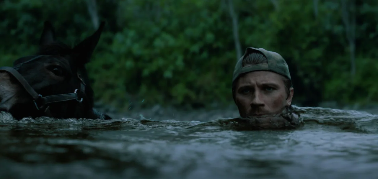 Triple Frontier (2019) movie still: medium — A man and a dark horse swim side-by-side through a river in the wilderness. Lush green foliage lines…