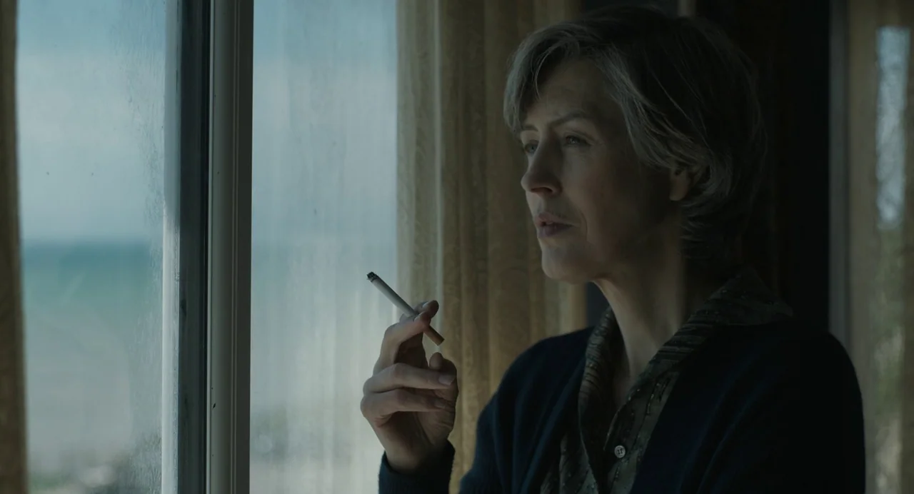 My Policeman (2022) movie still: closeup — A woman stands beside a large window looking out at an overcast coastal view. She holds a lit cigarette…