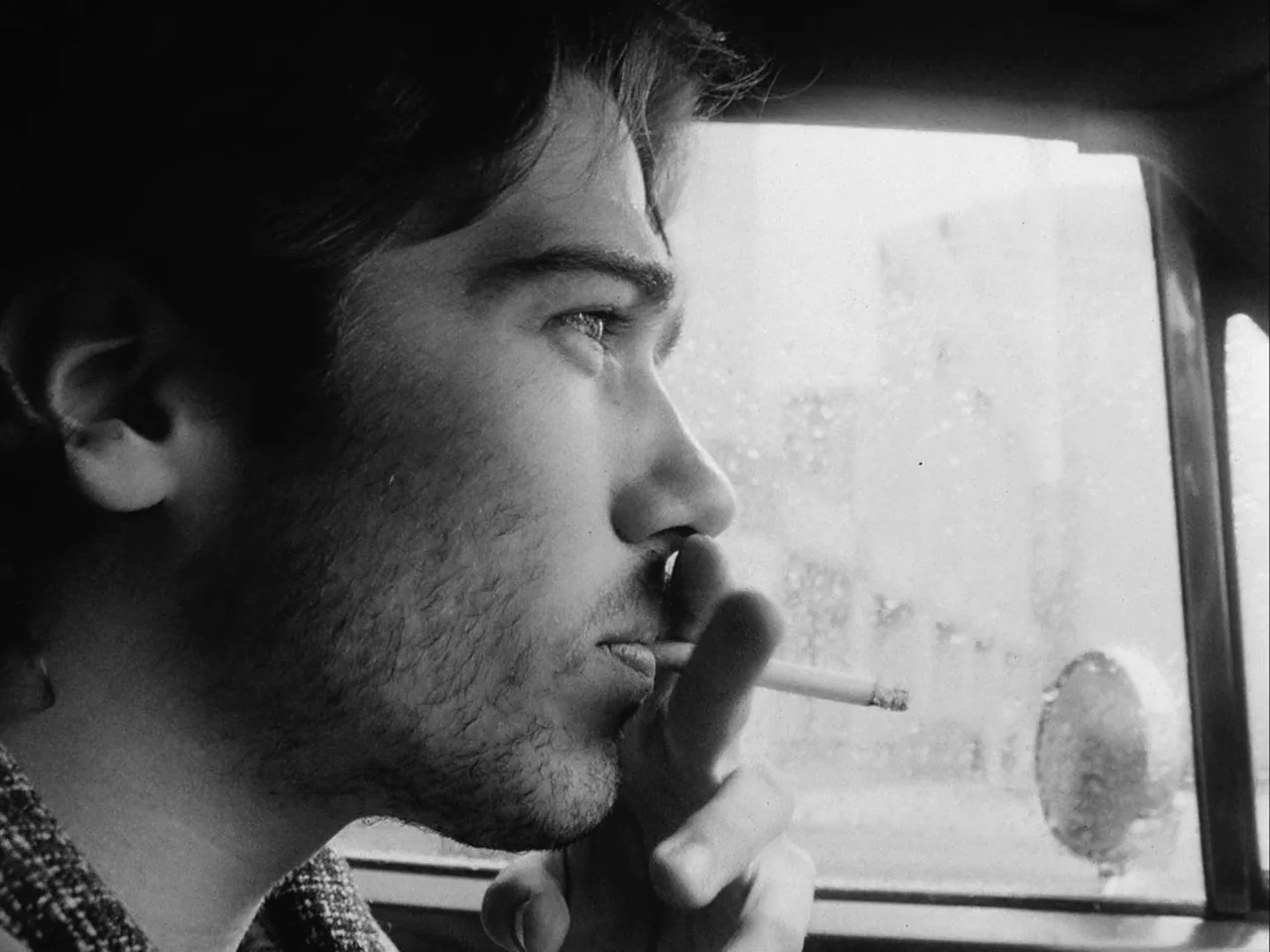 Mala Noche (1986) movie still: closeup — A man sits inside a vehicle looking through the rain-spattered side window. The interior of the car is dark…