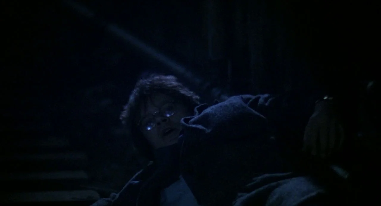 Jacob’s Ladder (1990) movie still: closeup — A person lies on the ground in a dimly lit, dark environment. The figure looks toward the camera with…